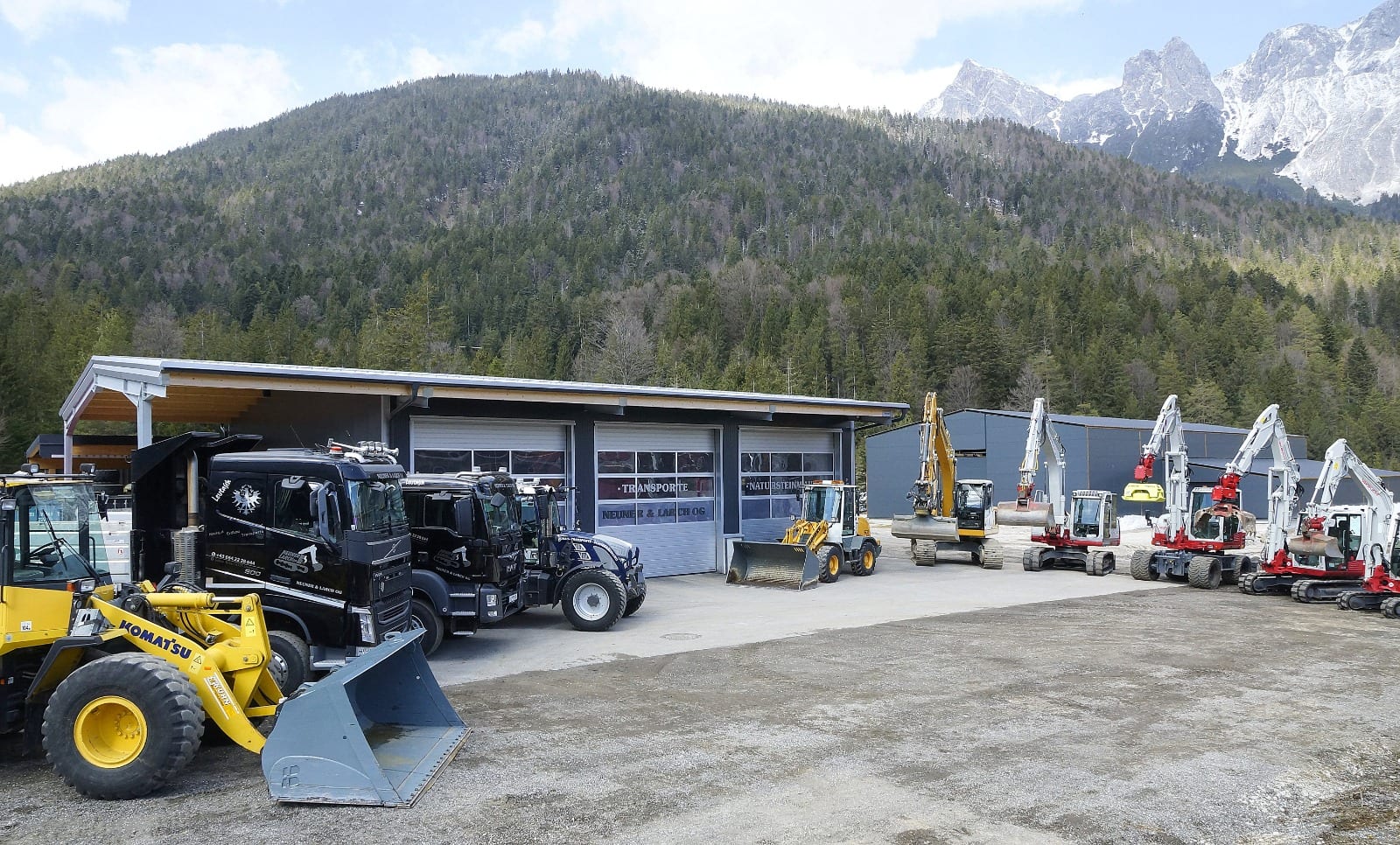 Panorama des Betriebsgeländes von Neuner & Larch mit Lastwagen, Radlader und einer Reihe Bagger vor bewaldeten Hängen.