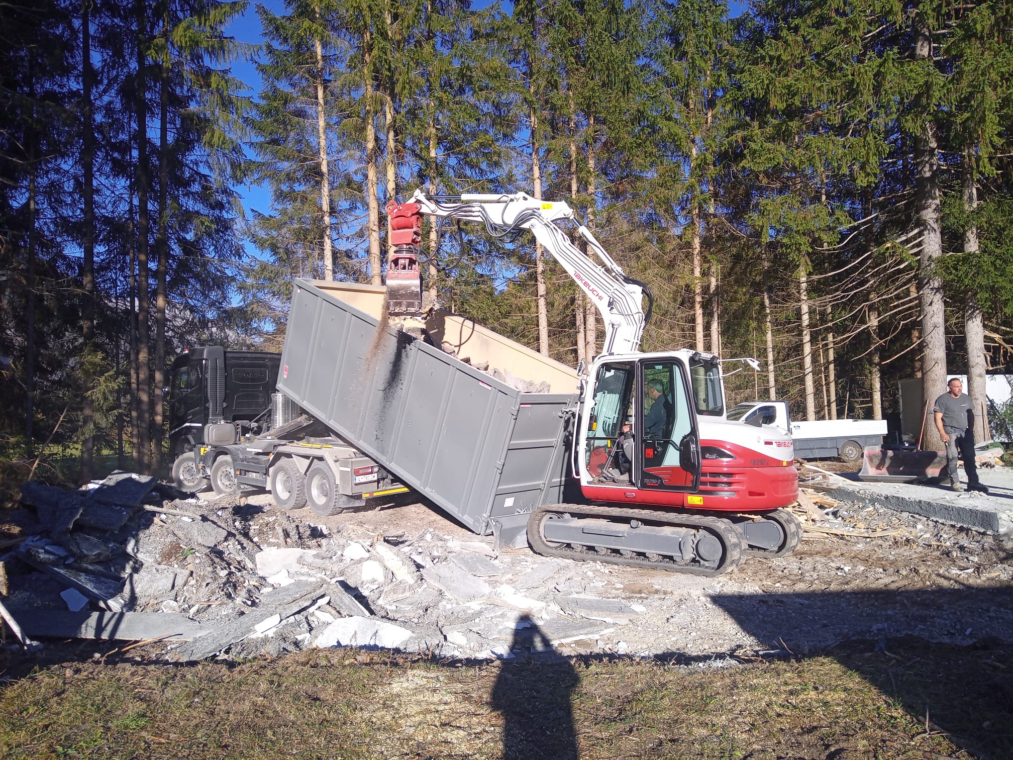 Ein Takeuchi-Bagger mit Abbruchgreifer belädt einen grauen Absetzcontainer auf einem Anhänger mit Bauschutt.