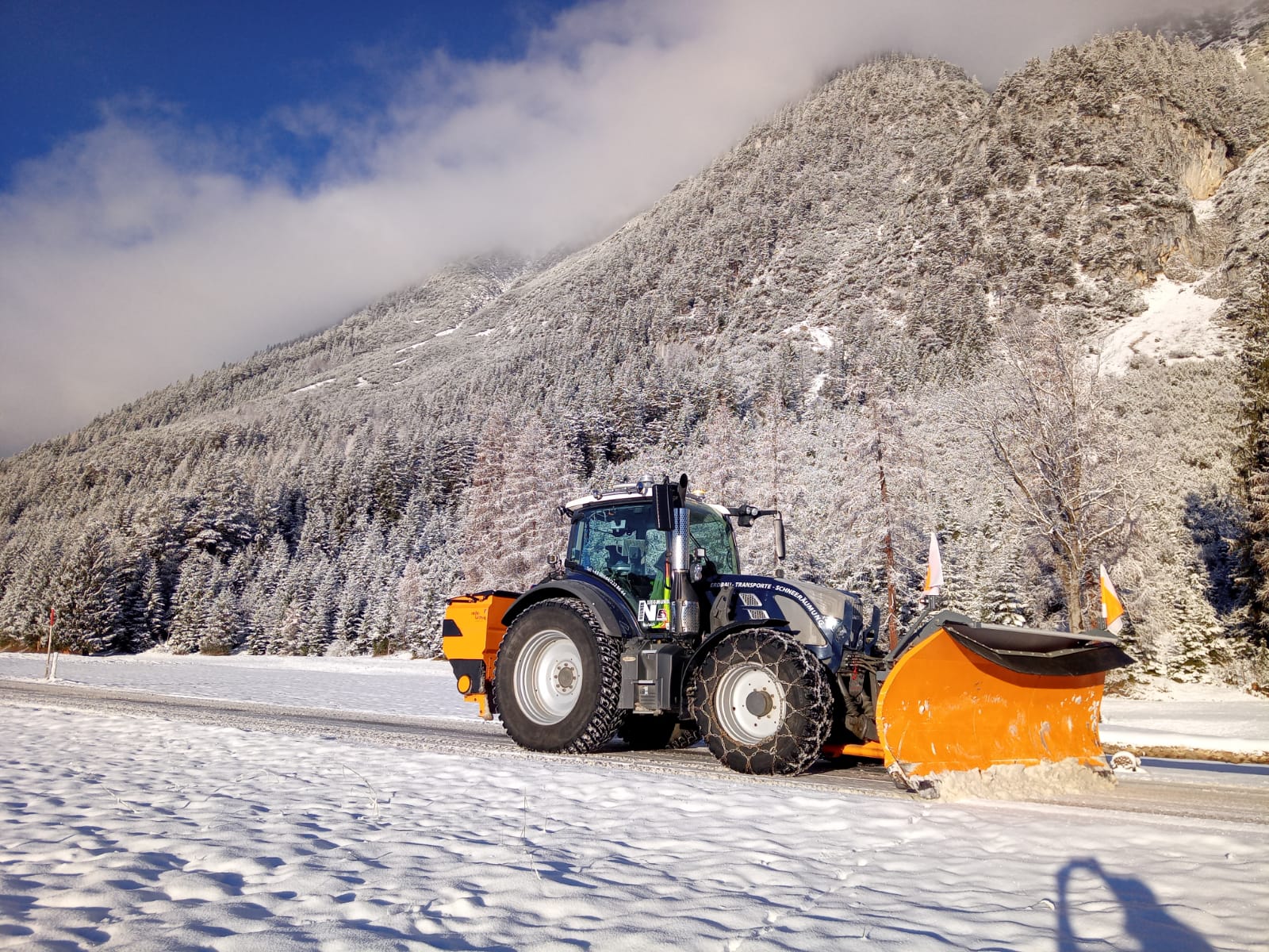 Traktor mit orangem Schneepflug räumt eine verschneite Bergstraße.